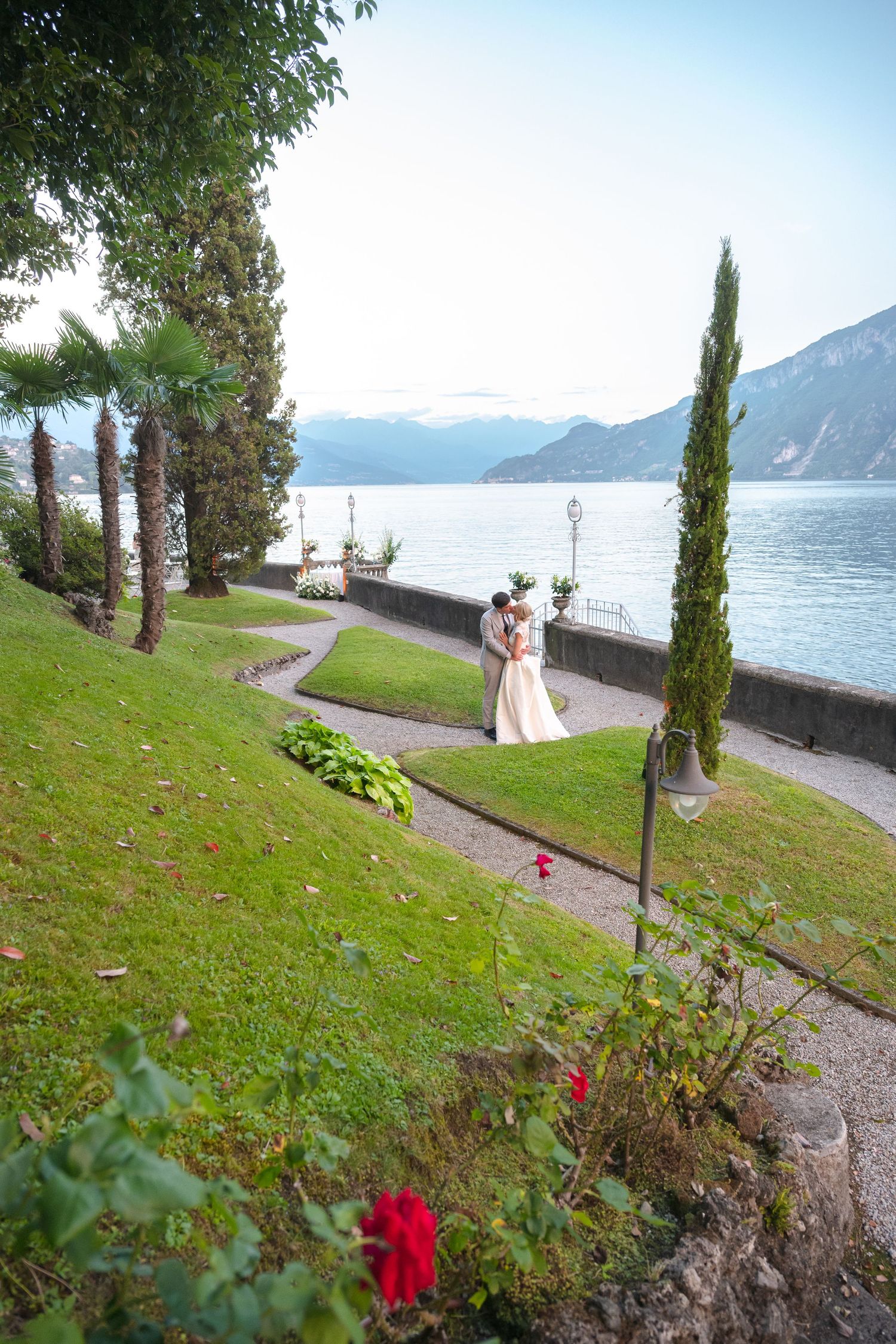A scenic lakeside pathway with cypress trees and red roses along a stone wall overlooking mountains and water.