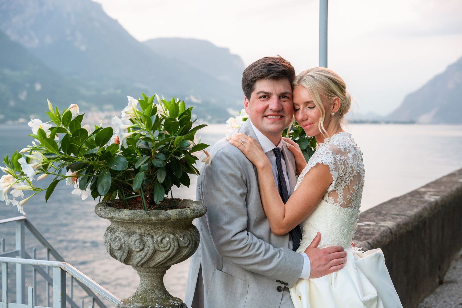 A romantic wedding couple embraces by a scenic mountain lake with lush greenery and misty peaks in the background.