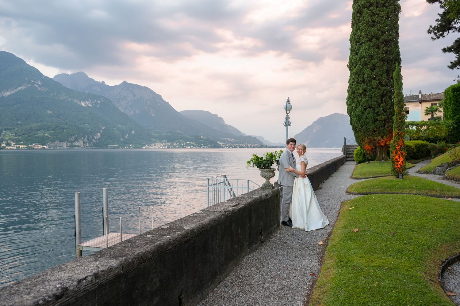 A scenic wedding photo at Lake Como, Italy with mountains and cypress trees along a lakeside stone wall.