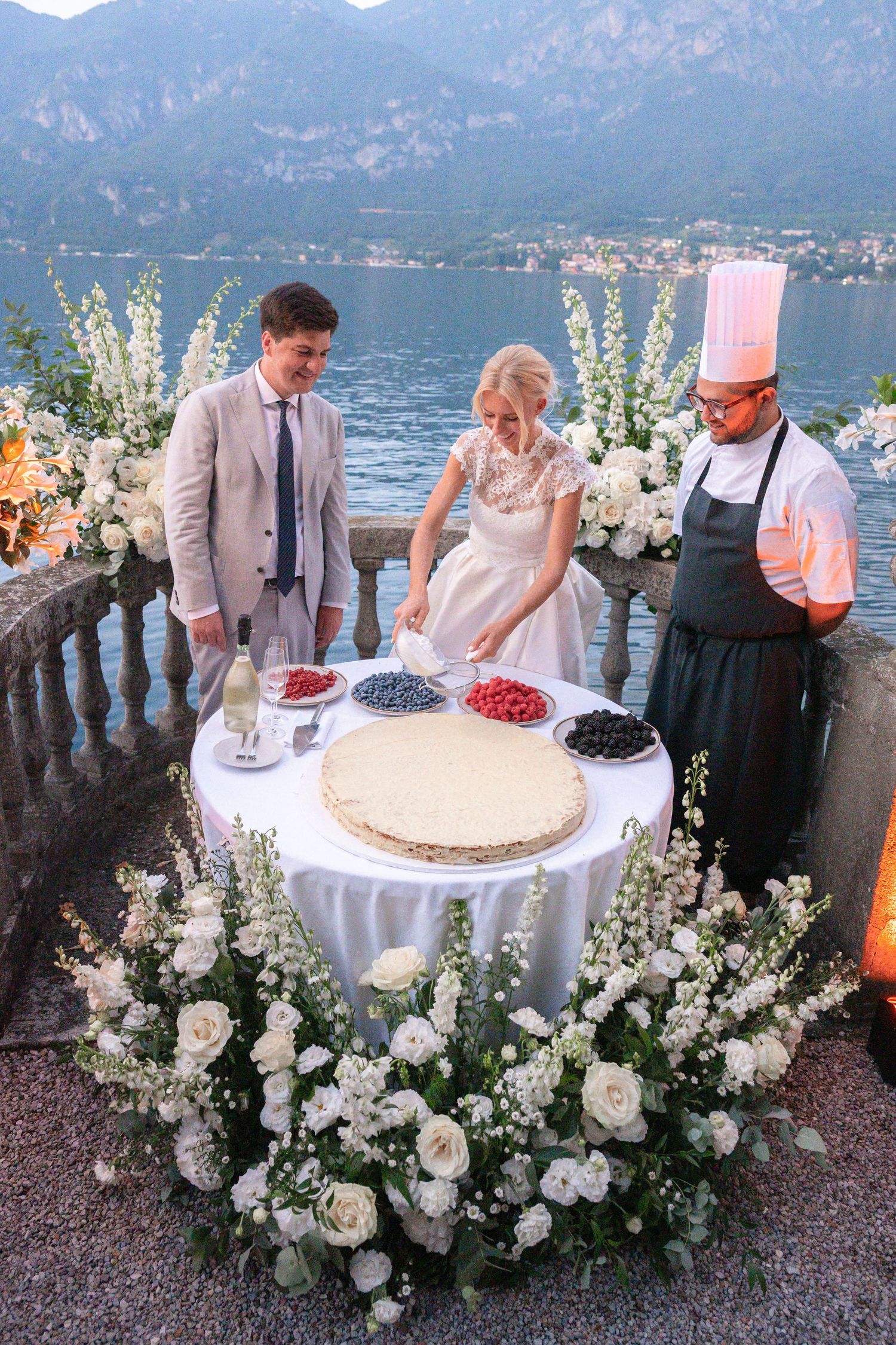 Wedding couple and chef prepare dessert display with fresh berries on lakeside terrace with mountain views and white flowers.