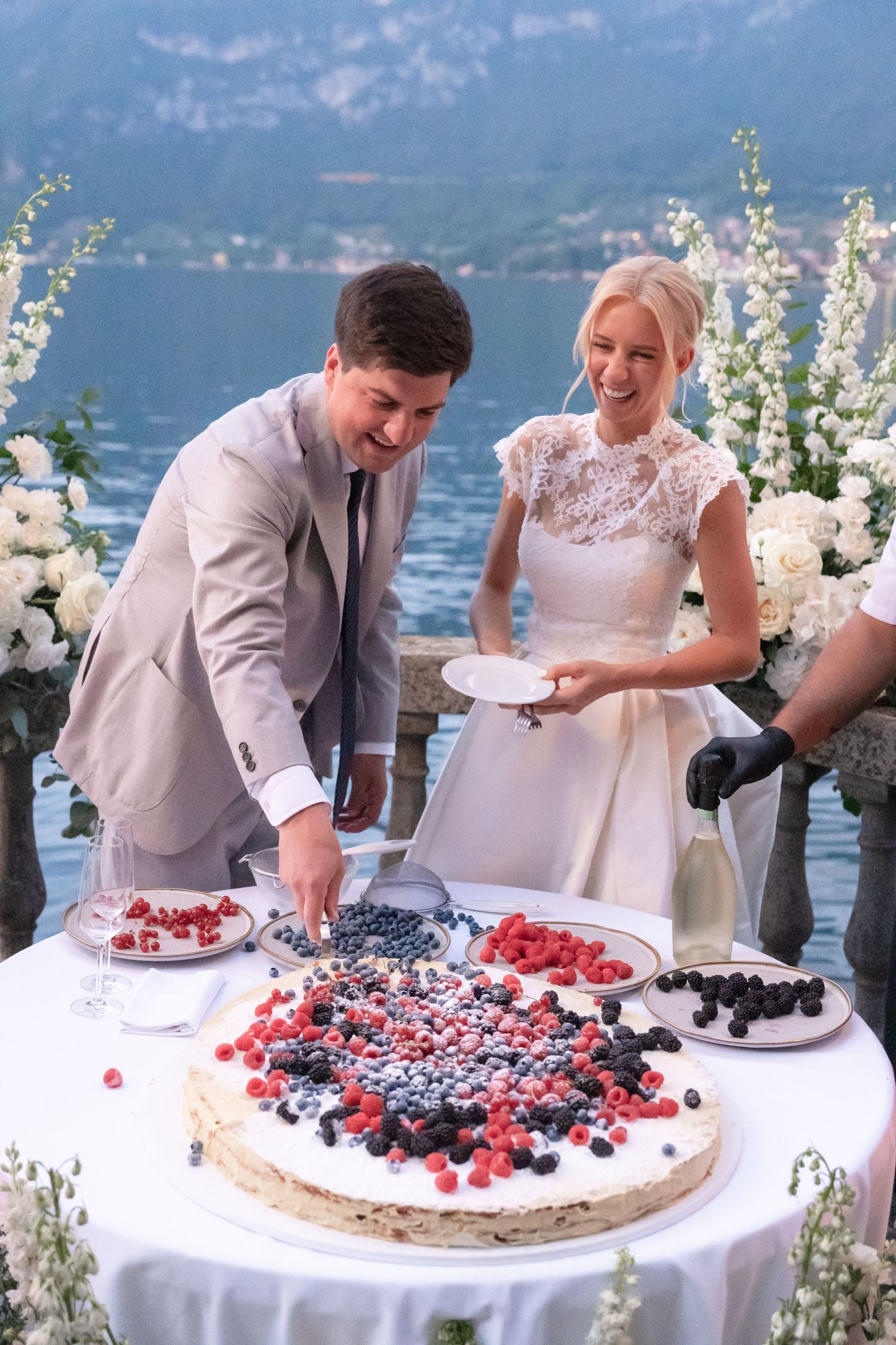 A wedding couple cuts and serves a rustic berry-topped cake by a scenic lakeside setting with mountains in the background.