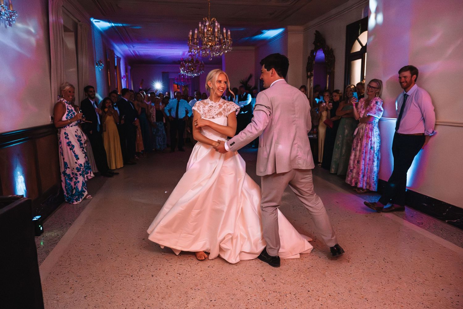 Wedding reception dance floor with couples dancing under purple lighting and chandeliers in an elegant ballroom setting.