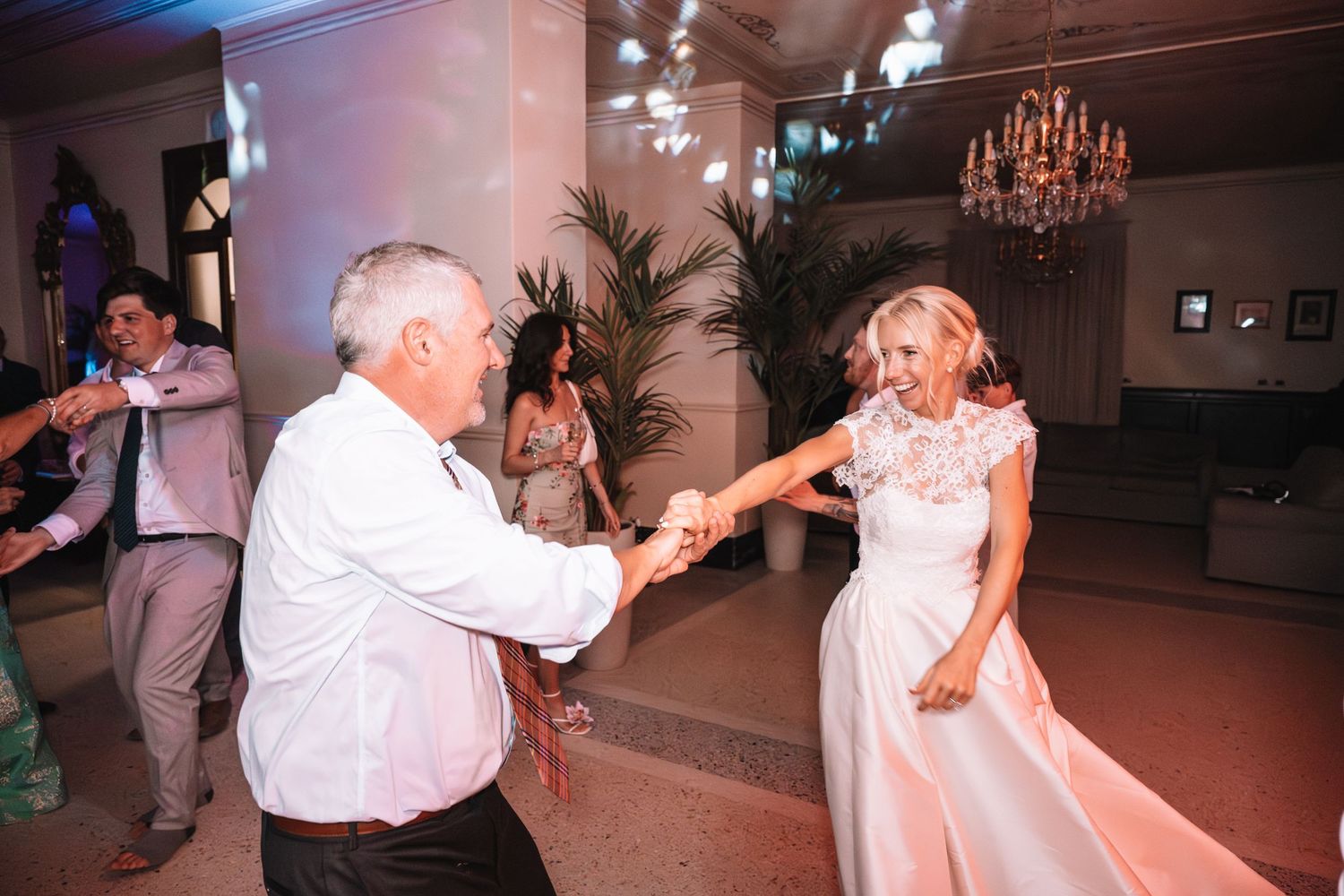 Wedding guests dance together in a dimly lit reception hall with chandeliers overhead.