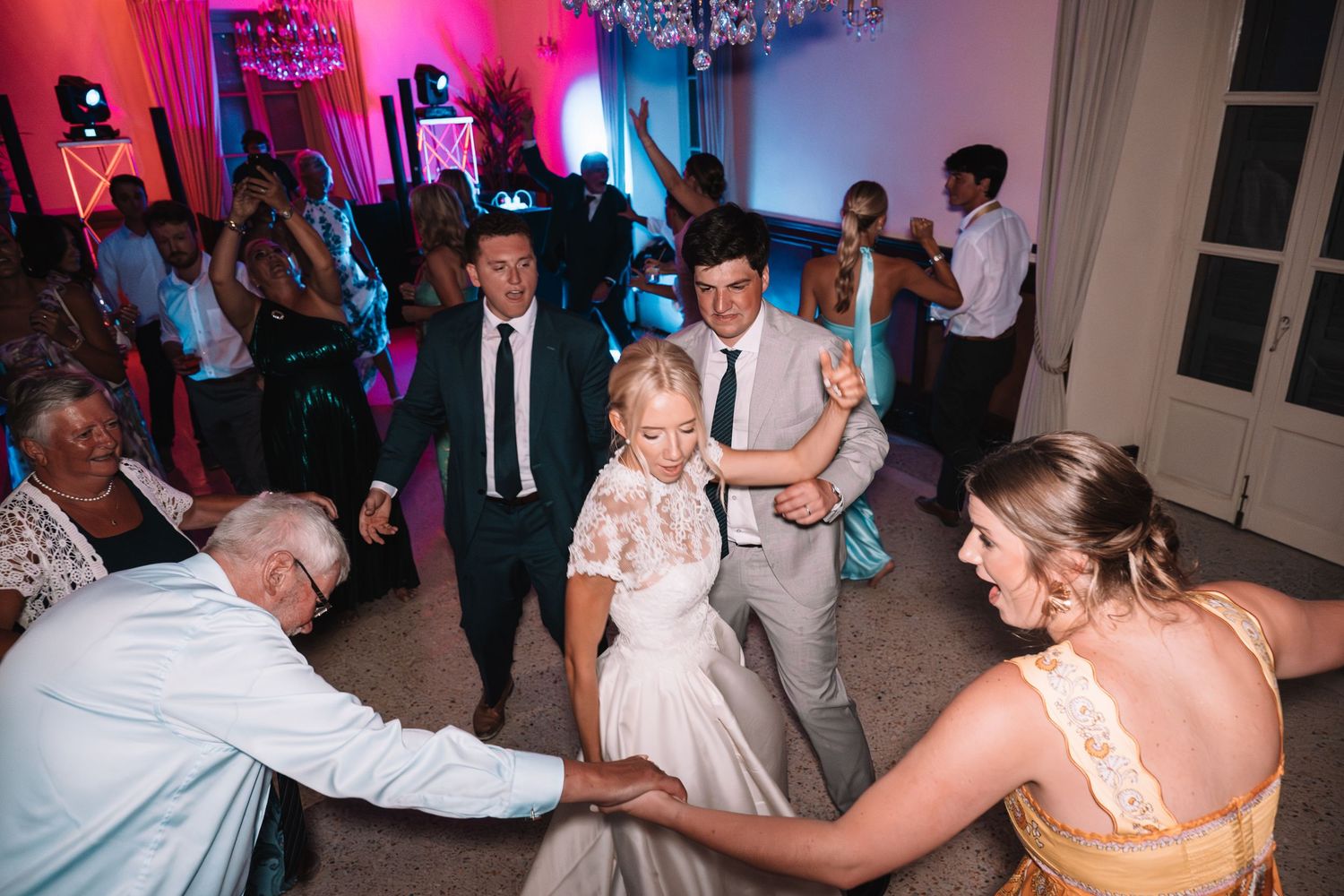 Wedding guests dance together on the dance floor illuminated by vibrant pink and blue lighting at an evening reception.