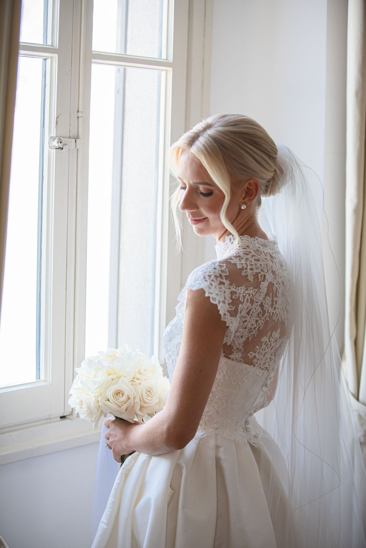A bride in a lace-detailed wedding gown with cap sleeves holds a white rose bouquet near sunlit windows.