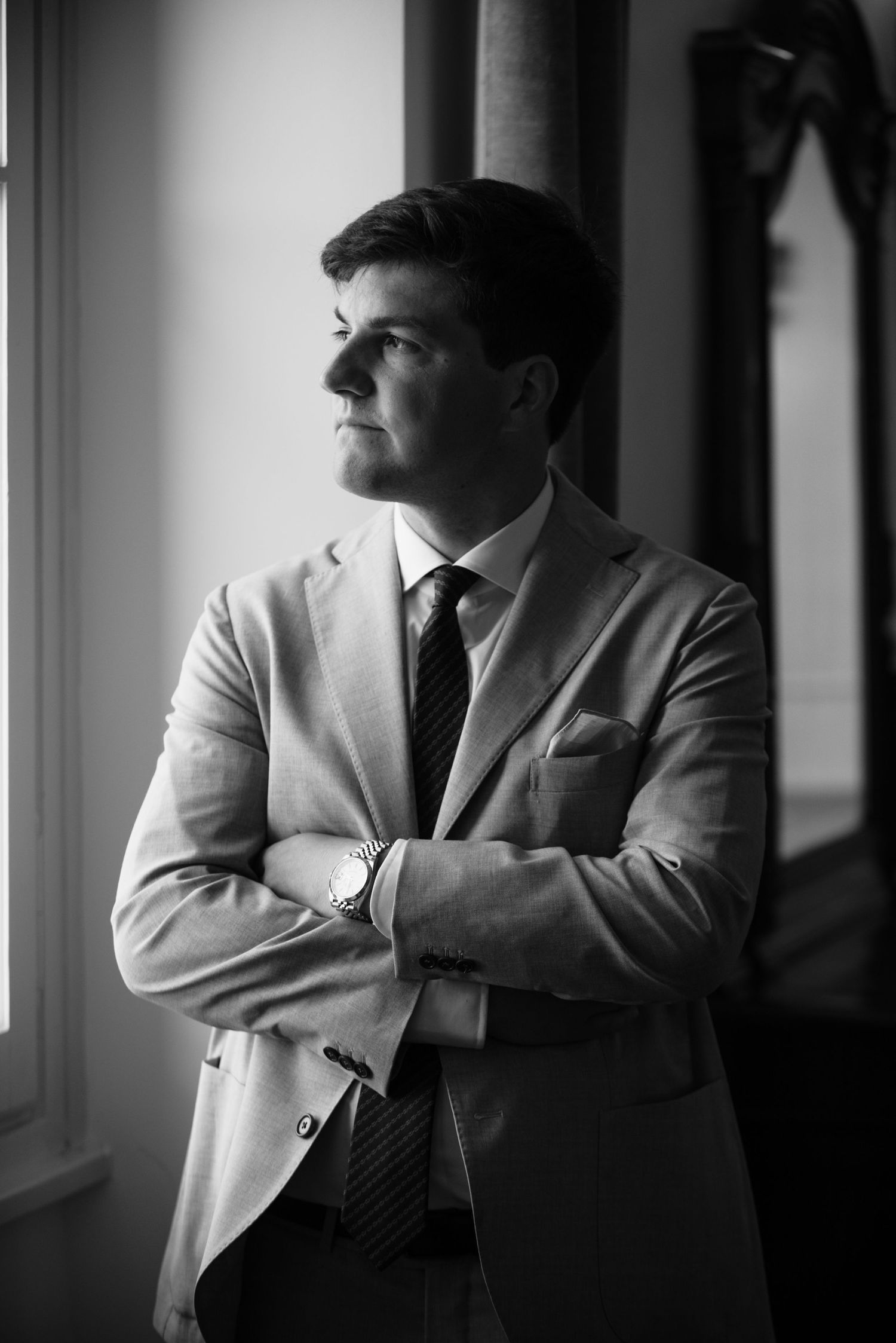 A person in a light suit jacket stands by a window in a moody black and white photograph series.