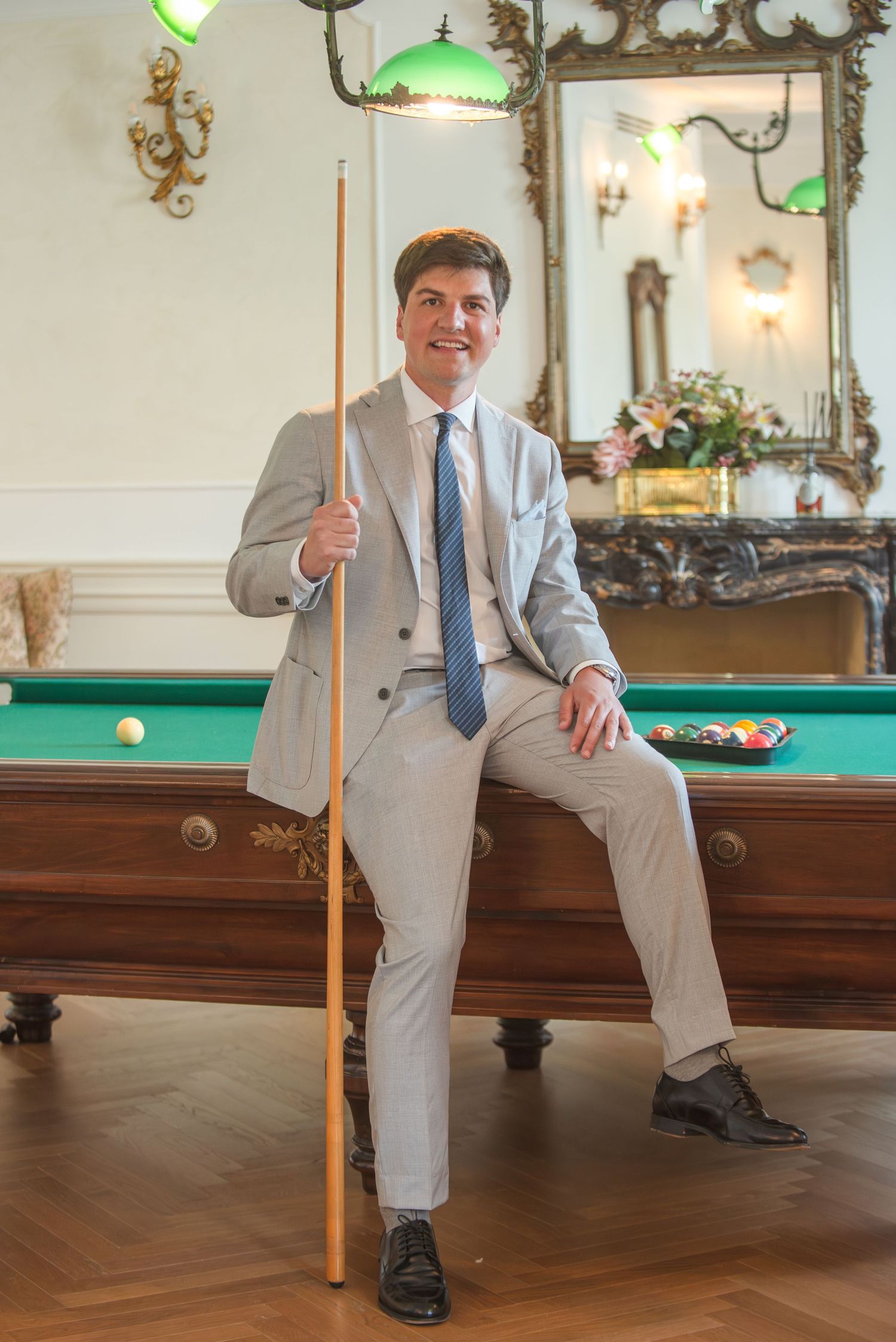 Someone in a light gray suit and blue tie sits casually on the edge of a vintage pool table in an elegant room.