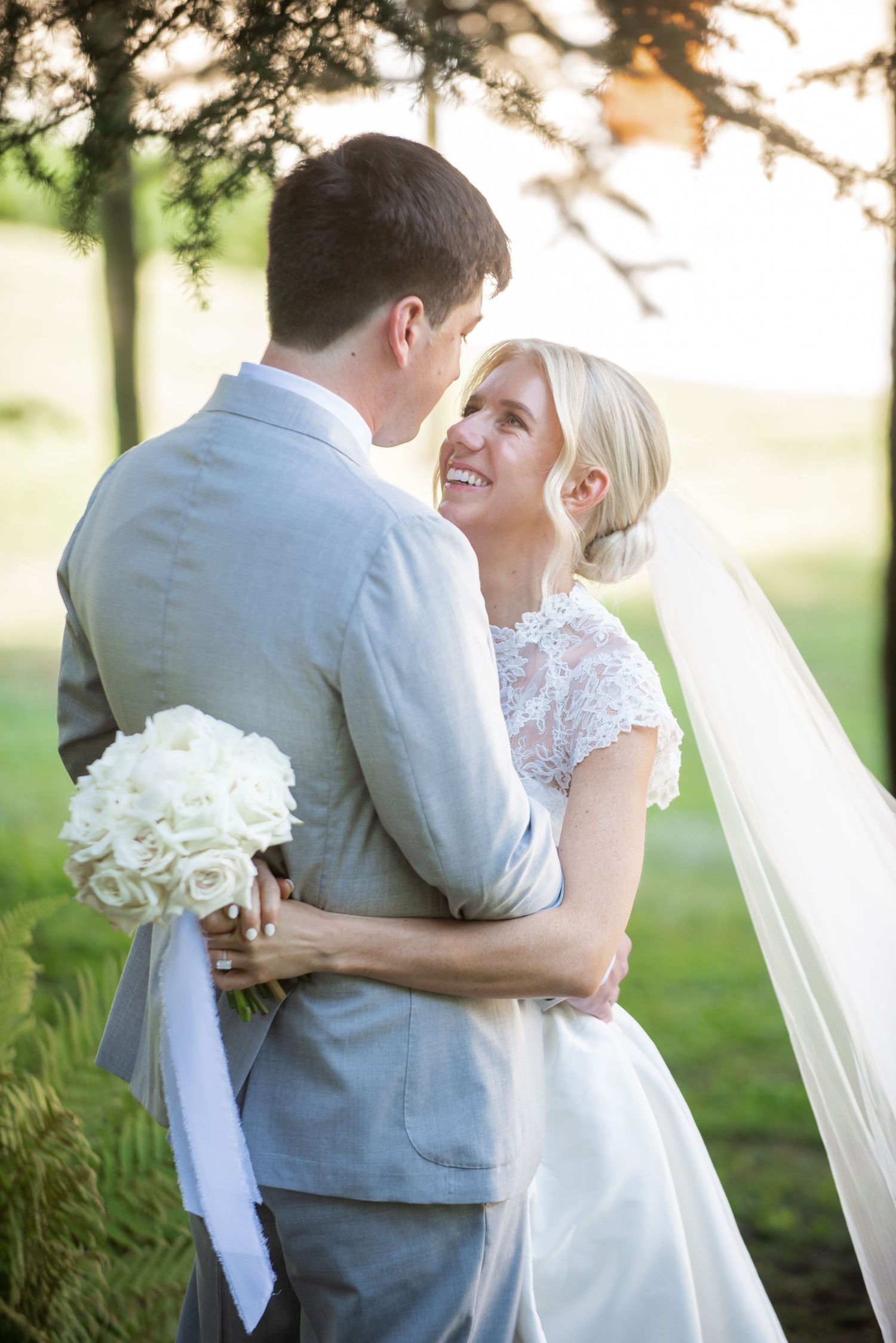 Romantic wedding couple embraces in sunlit garden setting wearing white dress and gray suit.