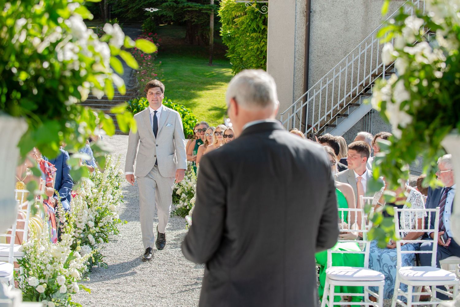 Wedding guests gather outdoors at a garden reception with white chairs and floral decorations on a sunny day.
