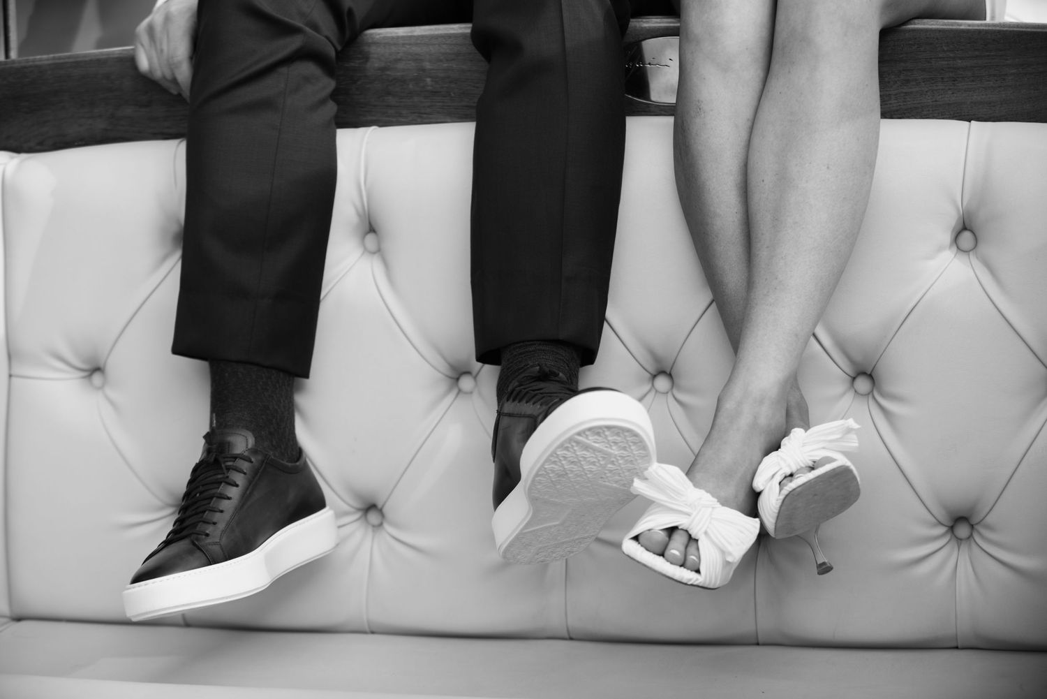 Black and white photo showing legs and feet of people sitting on a tufted leather bench or couch.