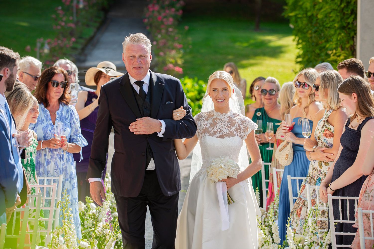 Wedding guests celebrate outdoors at a sunny garden reception with white chairs and decorative elements.