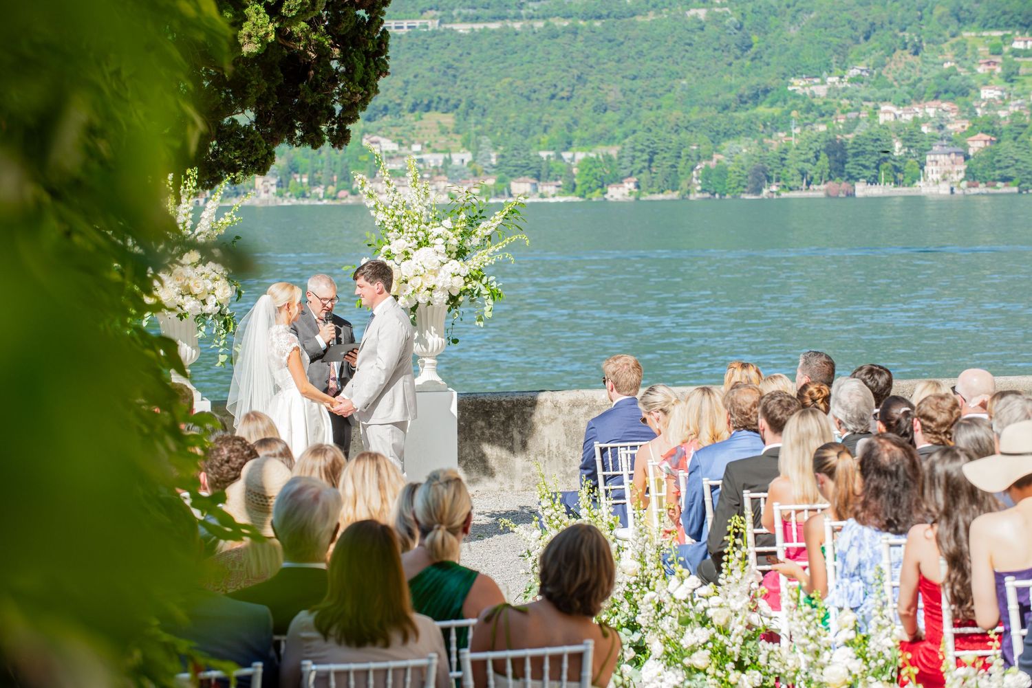 Guests gather for an outdoor lakeside wedding ceremony with white floral arrangements and mountain views in the background.