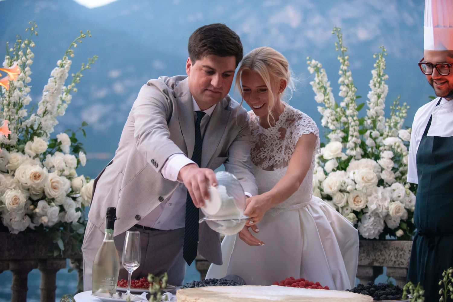 Couple shares a celebratory moment cutting their wedding cake together with white floral arrangements in the background.