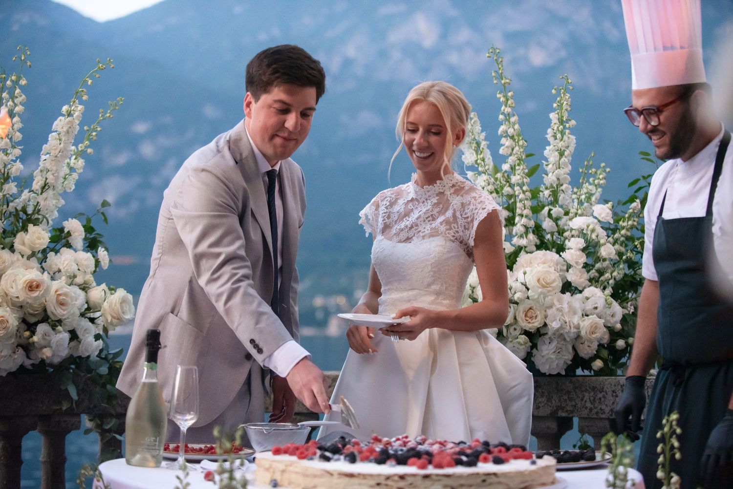 A series of wedding reception photos showing cake cutting, toasts, and decorative white floral arrangements by the lake.