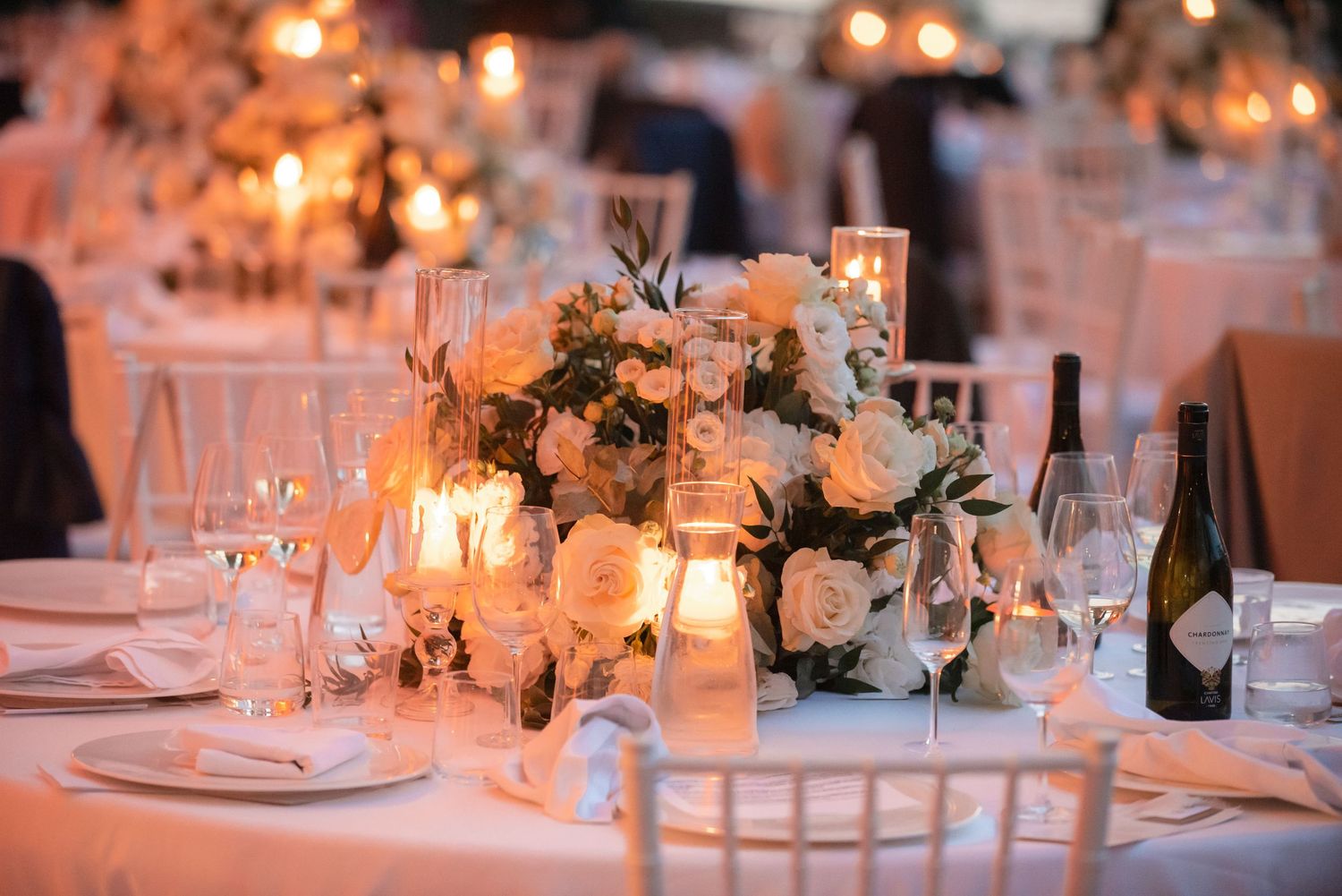 Elegant wedding reception table with white floral centerpiece and glowing candles on crisp white linens.