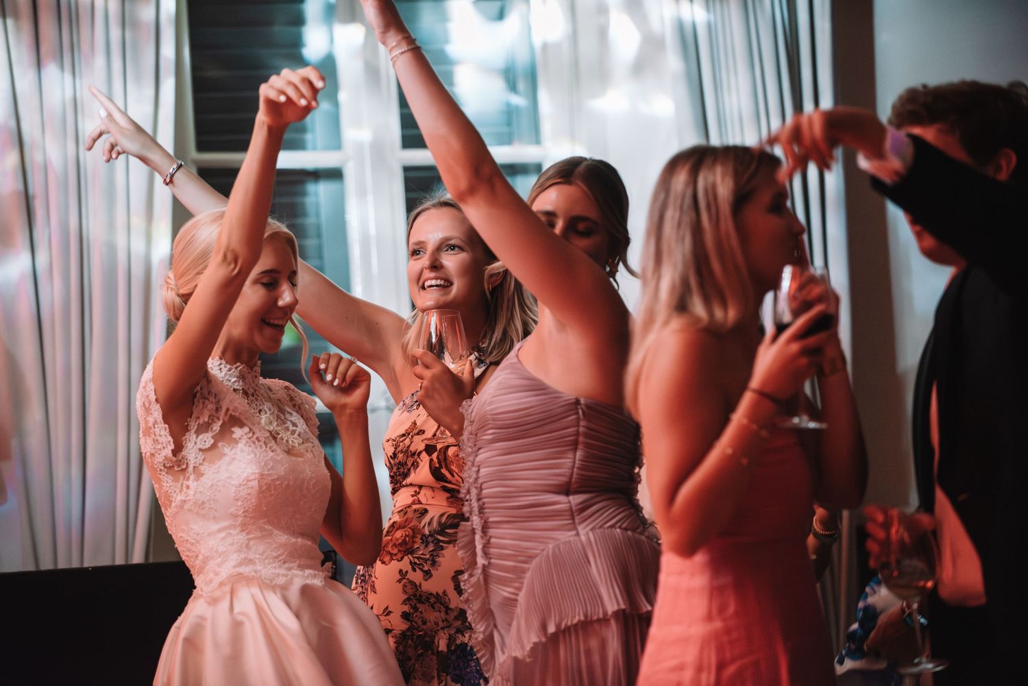 Joyful party guests in pink dresses dance and celebrate with raised arms at an indoor celebration.