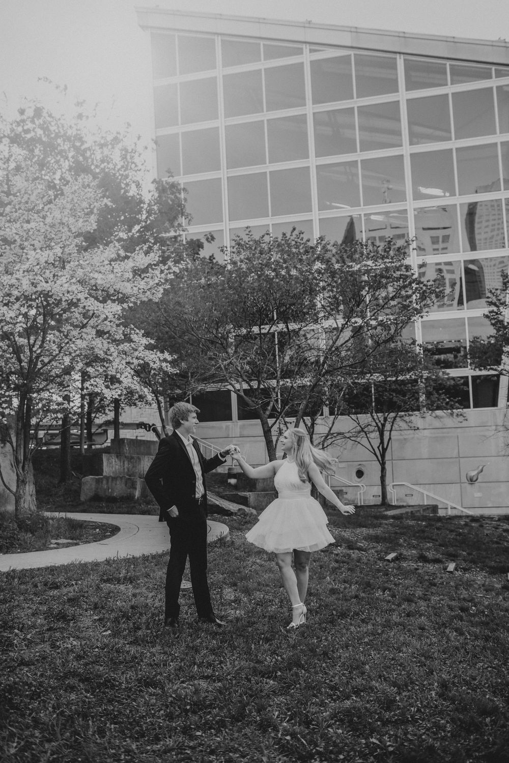 A couple in formal attire stands on a grassy lawn outside a modern glass building in black and white photography.