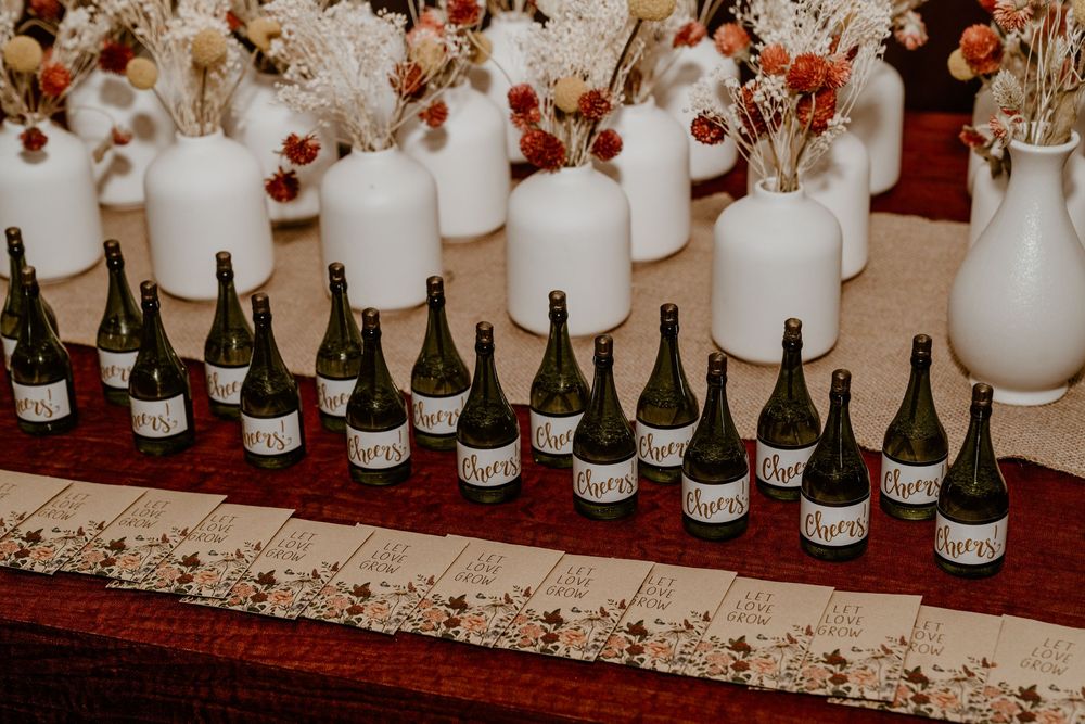 Green wine bottles with white labels arranged in a row on a burgundy table runner, with white vases and dried floral arrangements behind.