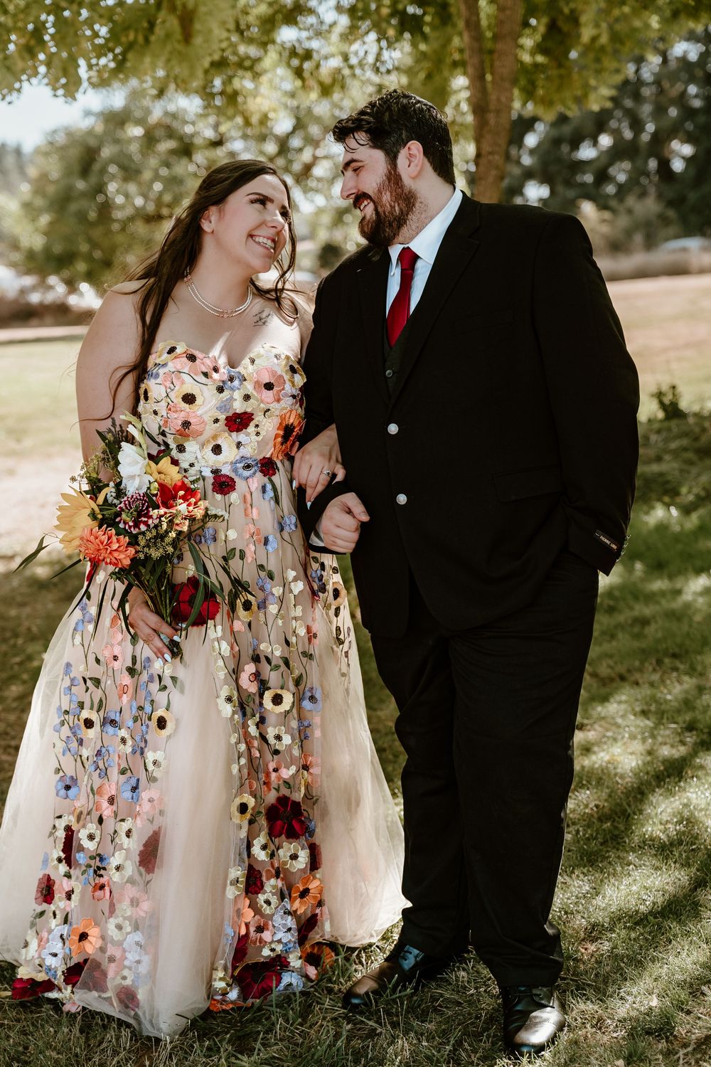 A bride in a floral gown and groom in a black suit share a loving glance outdoors on their wedding day.
