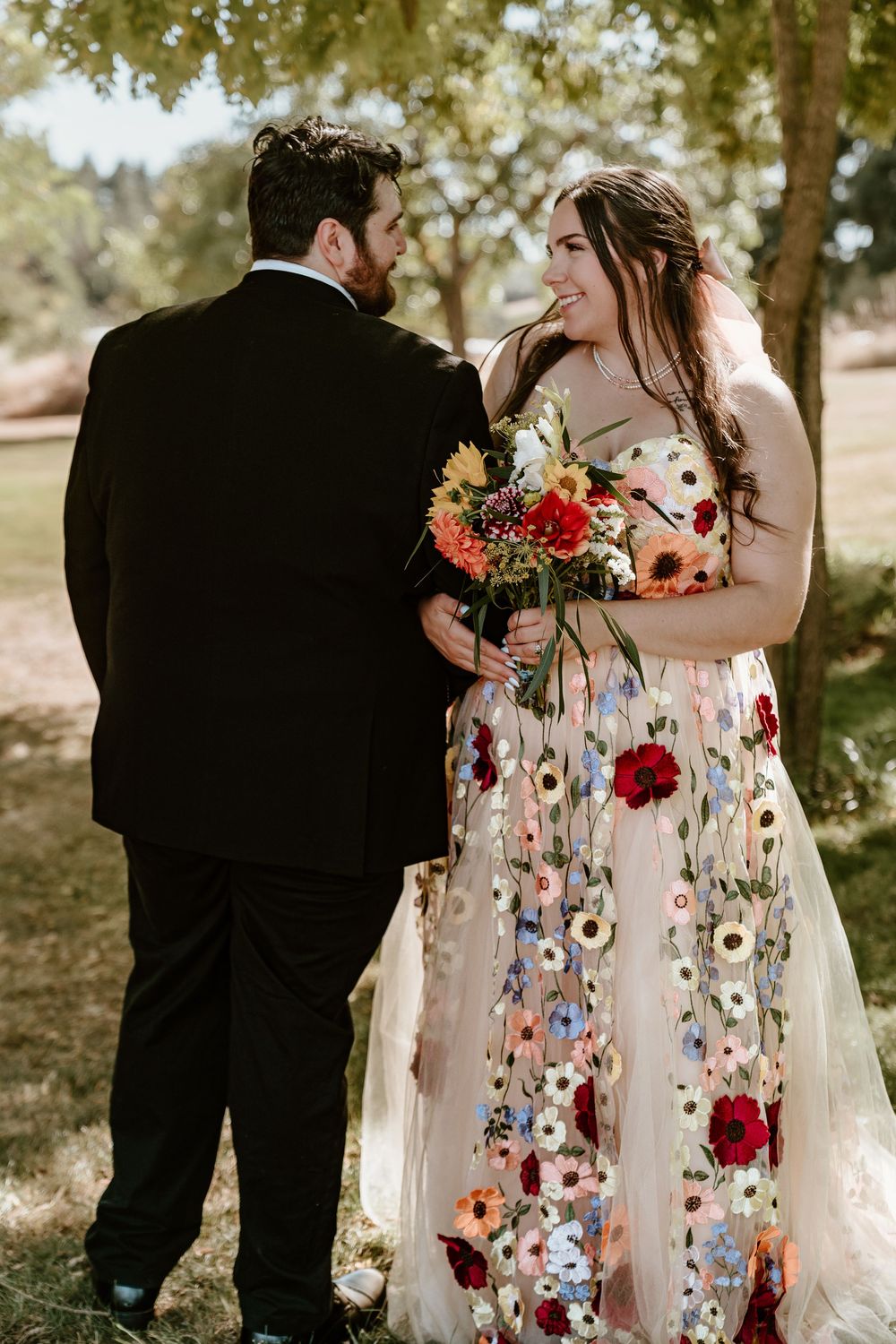 Bride in floral embroidered wedding gown holding colorful bouquet stands with groom in black tuxedo outdoors.