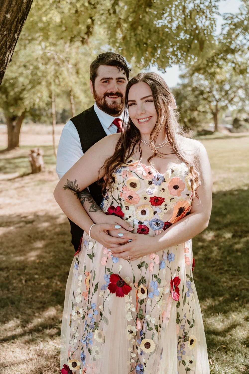 A smiling couple poses outdoors, woman in floral gown, man in vest with red tie, embracing under green trees.