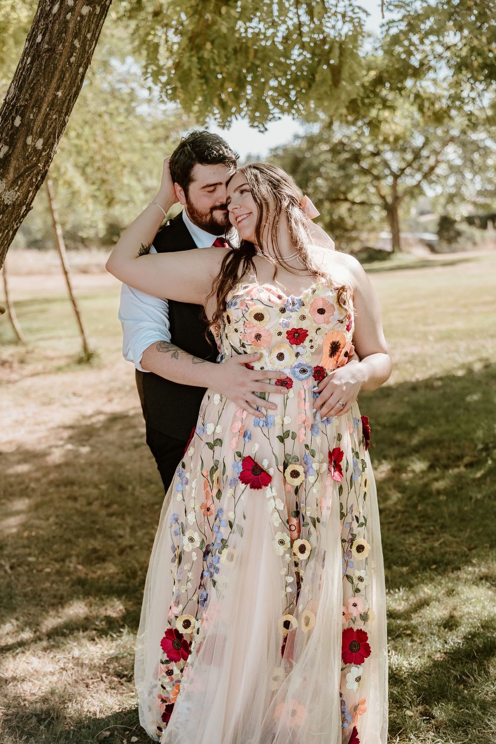 Couple embracing outdoors, woman in floral embroidered gown, man in dark suit, sharing romantic moment under trees.
