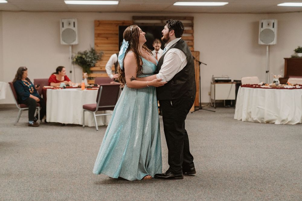 Couple shares first dance in teal wedding gown at elegant reception hall with guests seated at decorated tables.