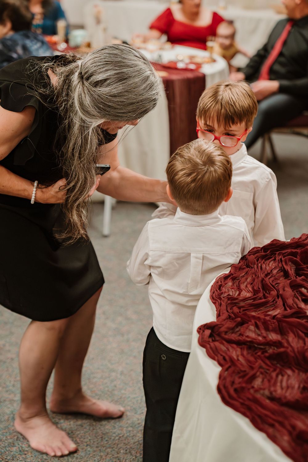 Woman in black dress bending down to speak with two young boys in white shirts at a wedding reception.