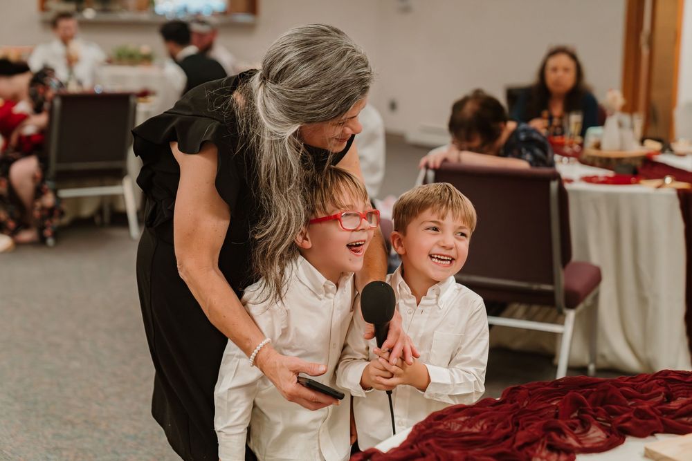Woman in black dress hugs two laughing boys in white shirts holding a microphone at a festive indoor event.