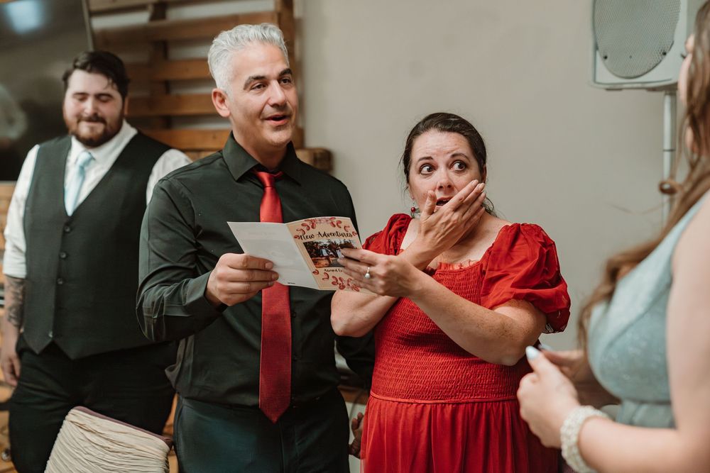 Man in red tie reads card as woman in red dress reacts with surprise at a wedding reception.
