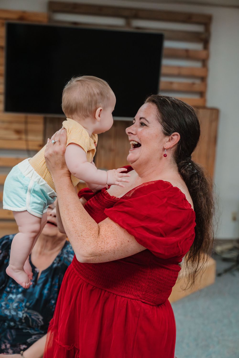 A joyful woman in a red dress holds a baby in a yellow outfit, laughing and sharing a tender moment indoors.