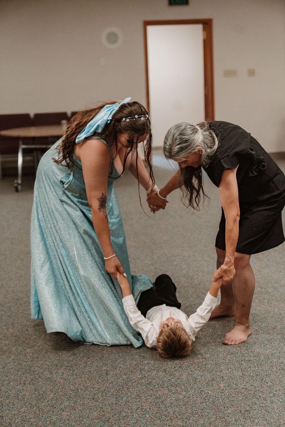 Two women in formal attire playfully hold a child lying on the floor at an indoor event.