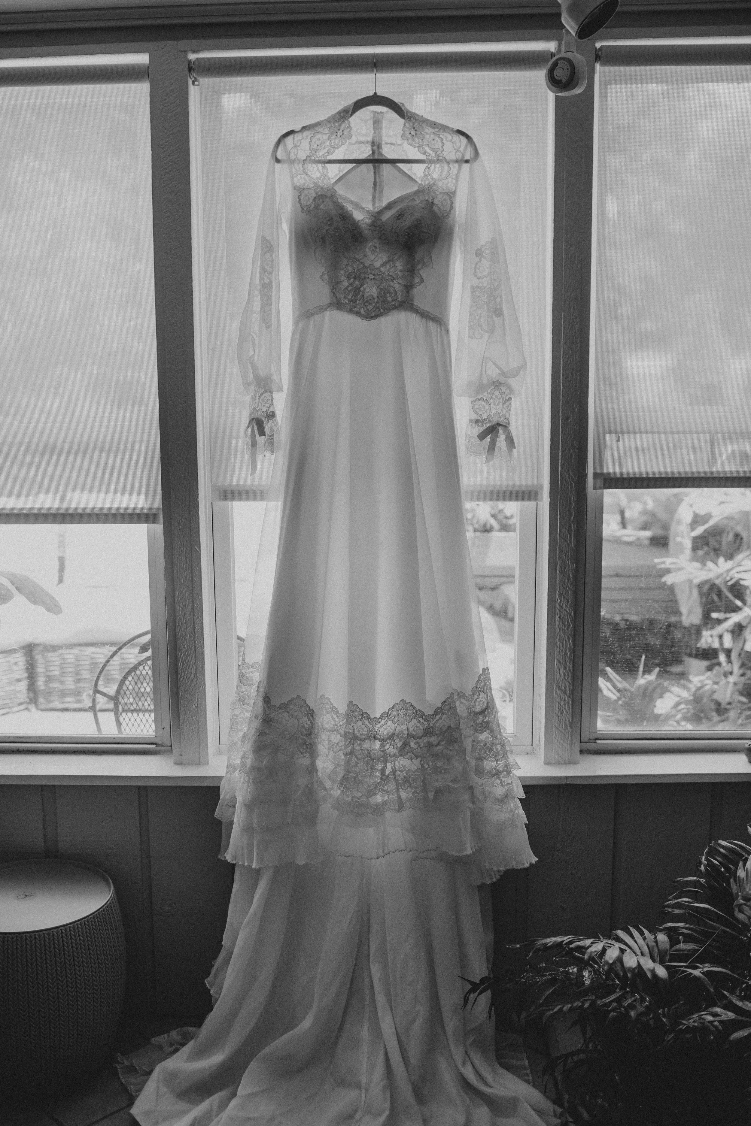 Vintage-style wedding dress hanging in window light displaying delicate lace details and flowing fabric.
