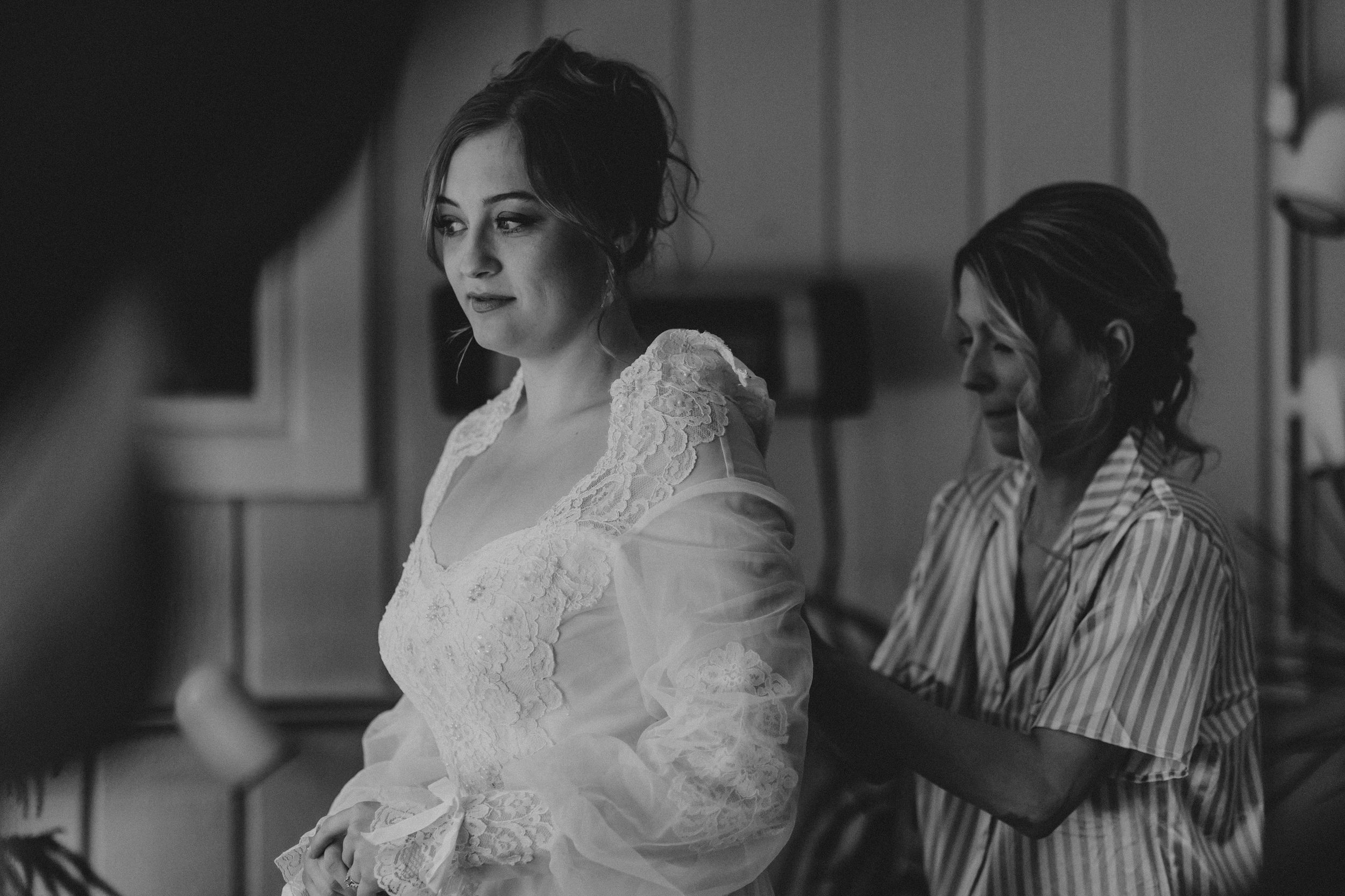 A black and white photo shows a bride getting ready in her lace wedding dress while an assistant helps behind her.