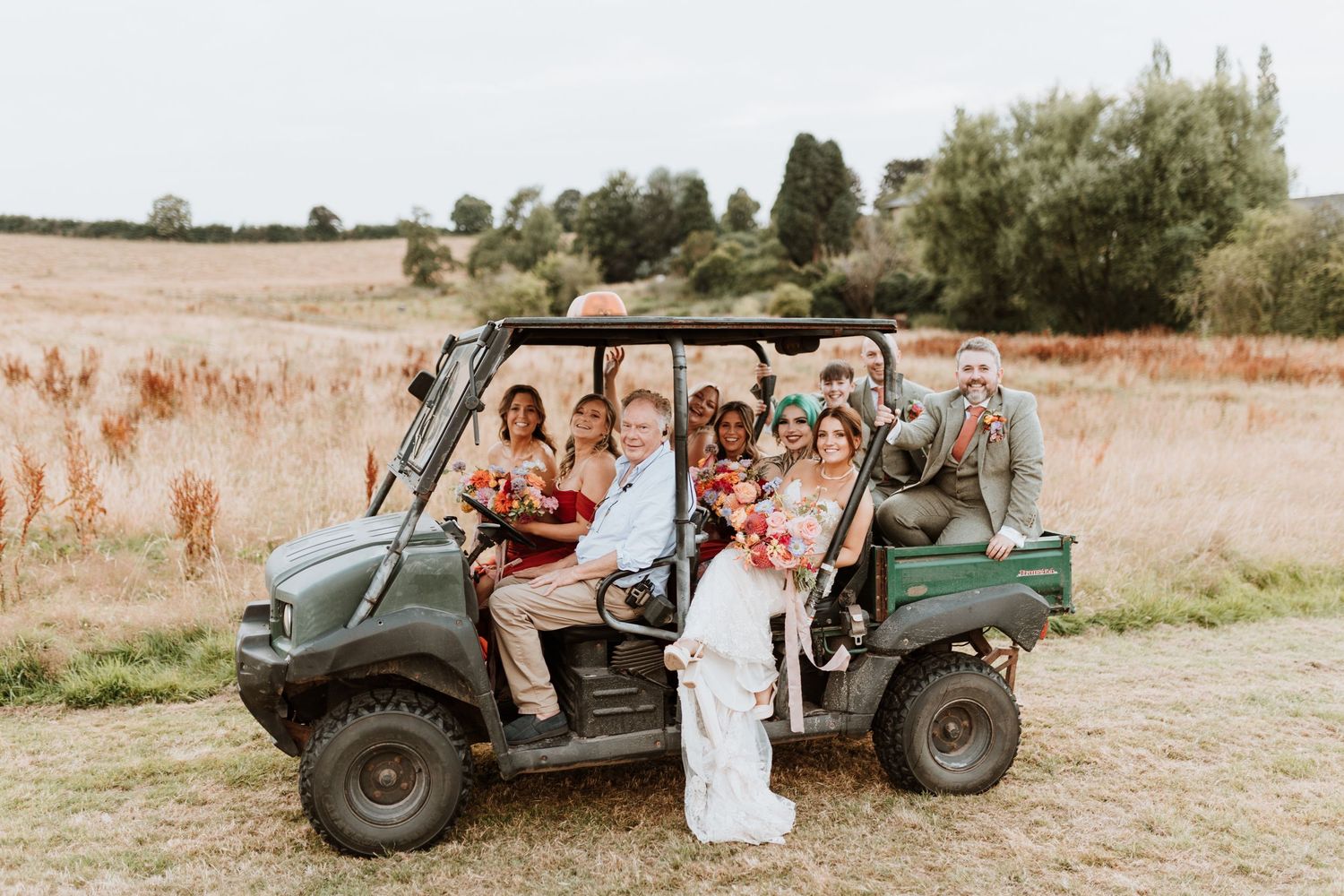 A vintage military-style jeep drives across a sun-drenched field with passengers in wedding attire.
