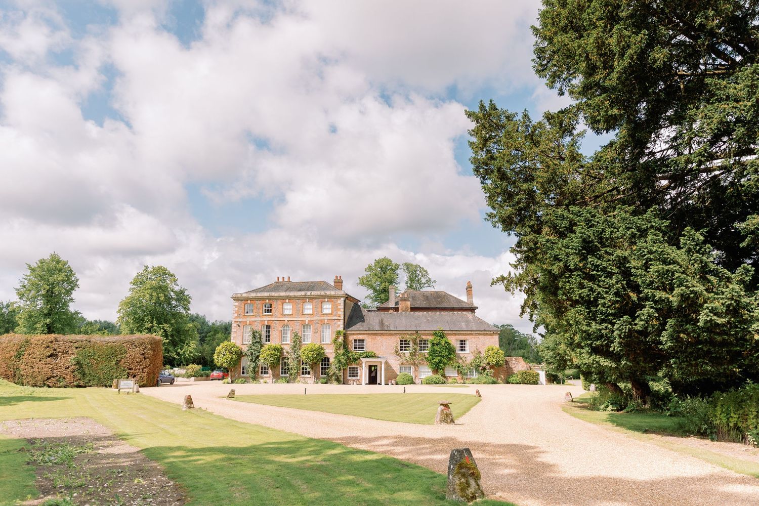 A grand country manor house with manicured gardens and gravel driveway sits under a bright summer sky with fluffy clouds.