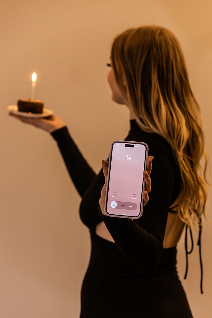 Blonde woman in black backless dress holding birthday cupcake with candle and showing phone screen.