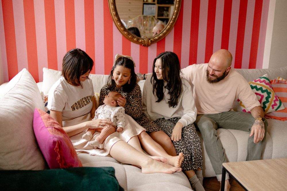 A group sits on a couch together looking at a small baby in a pink dress.
