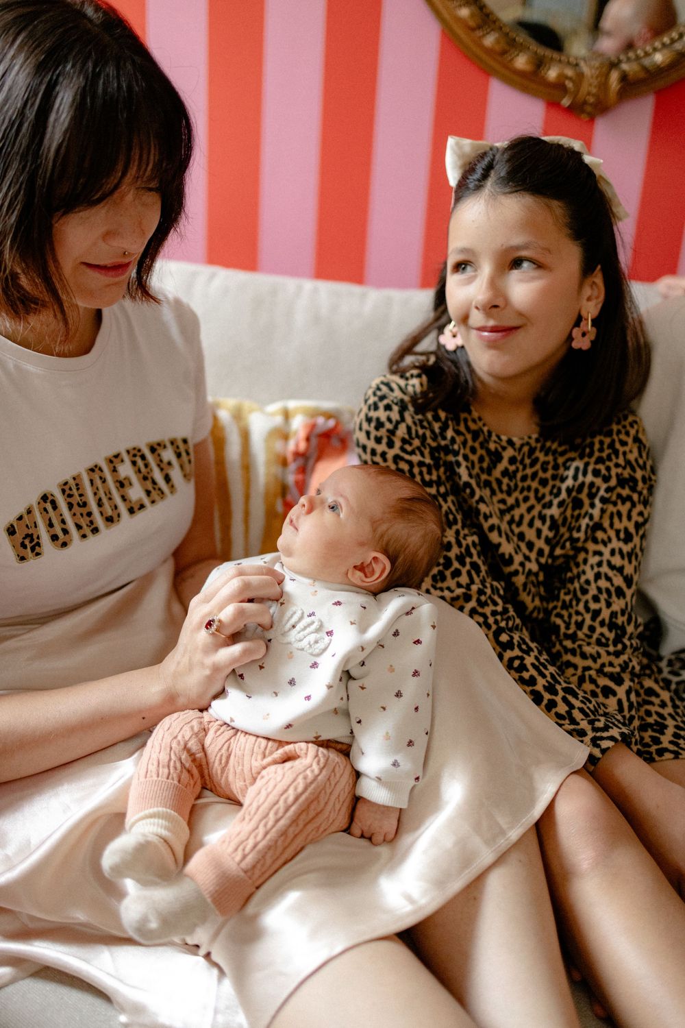 Parent in leopard print dress holds infant wearing white outfit against pink and white striped background.