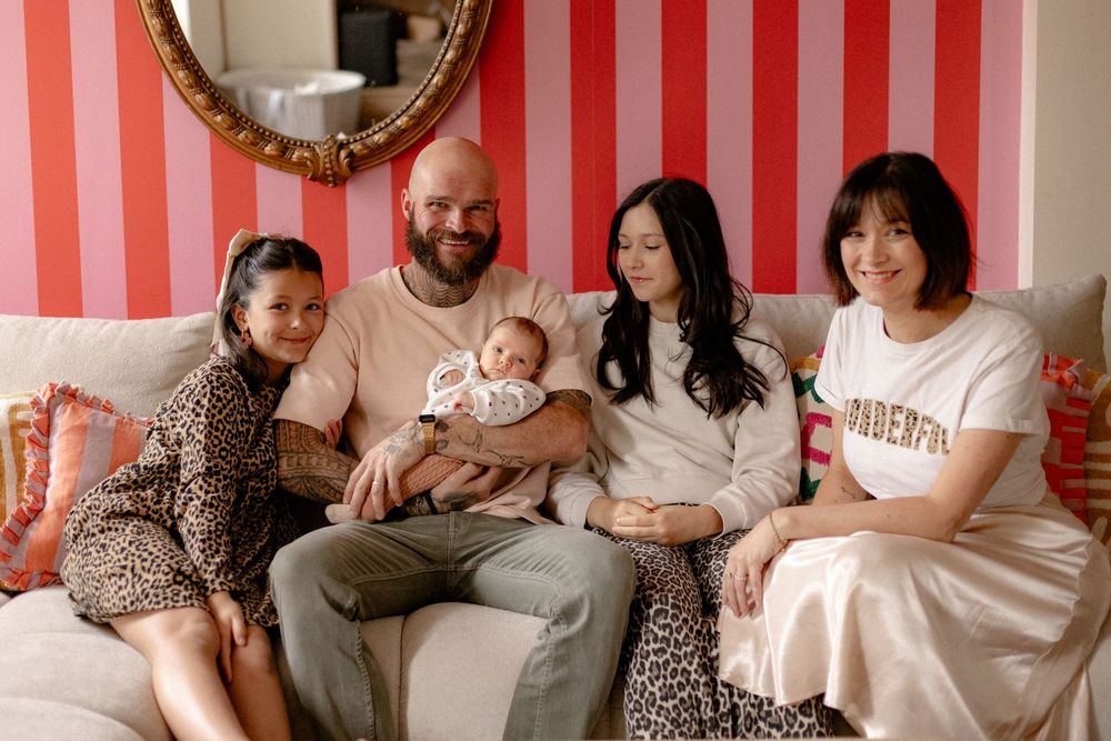 A family group sits together on a couch against a pink and white striped wall in a cozy indoor setting.