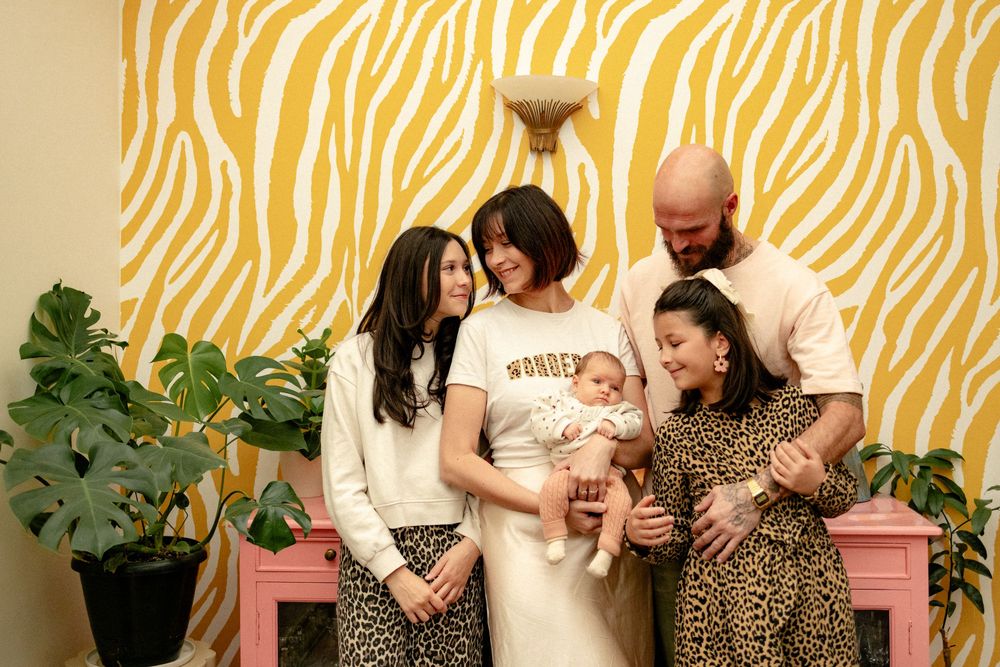 Family moment captured against yellow zebra print wallpaper with pink chair and houseplants.