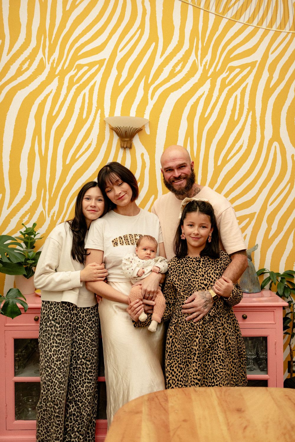 Family poses together against yellow zebra print wallpaper with pink bench and indoor plants.