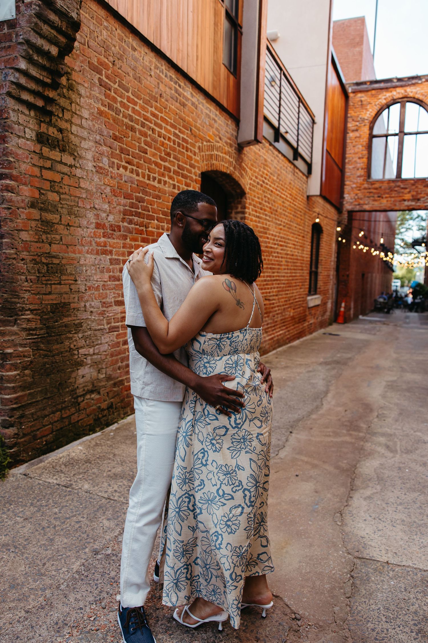 A couple embraces in a romantic moment against rustic brick walls in an alley with string lights visible in the background.
