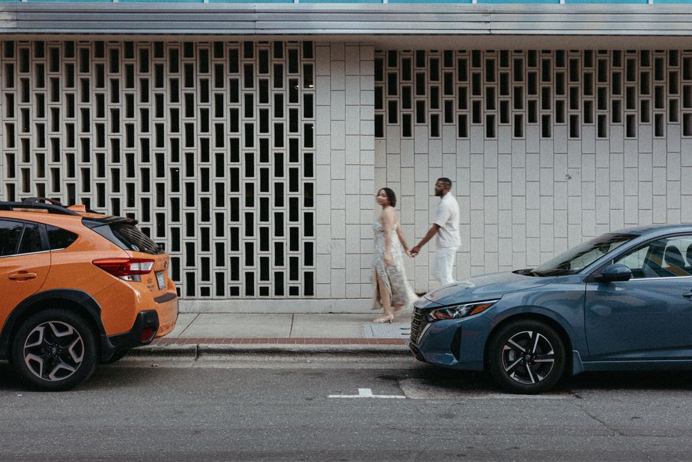 Two modern sports cars, one orange and one blue, parked side by side on a city street with a white wall backdrop.