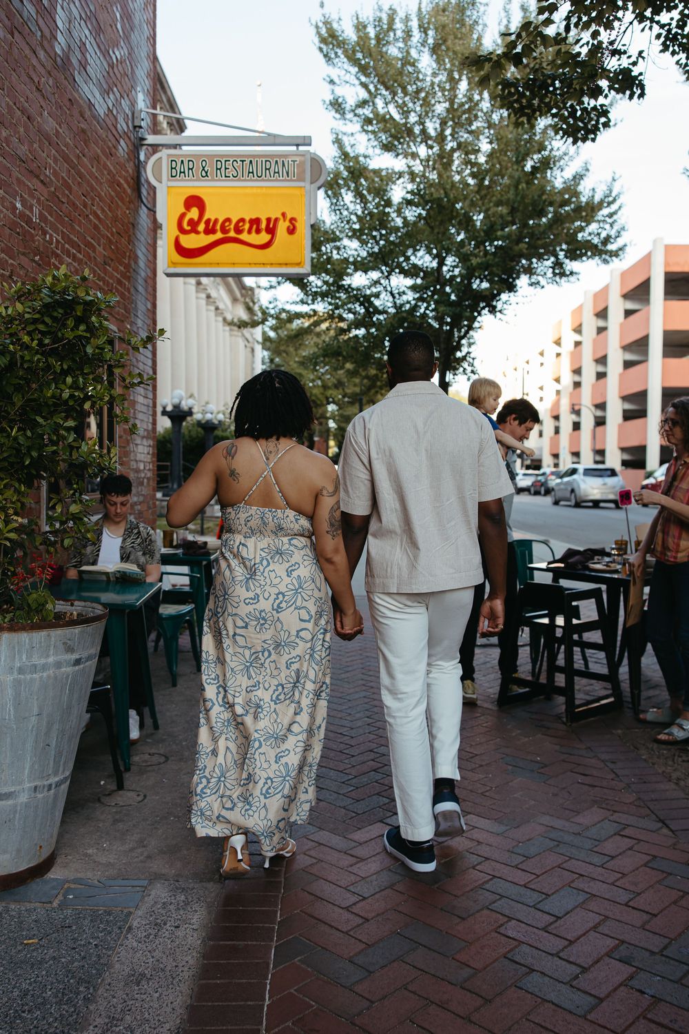 Two people walk together on a brick sidewalk past a restaurant sign on a sunny day.