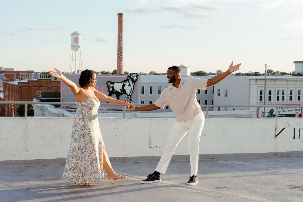 A couple in wedding attire dances and poses together on a rooftop with city buildings in the background.