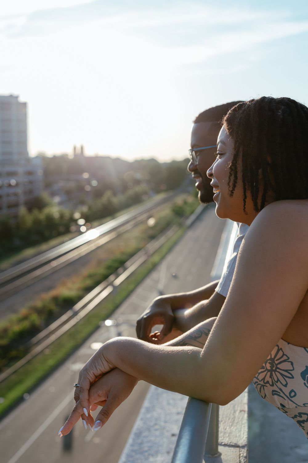 Two people lean on a balcony railing while enjoying a sunset view over an urban street on a warm evening.