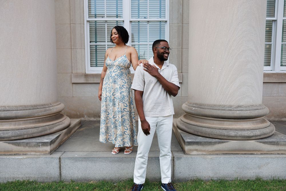 A couple in summer outfits poses together against a white stone column exterior of a building.