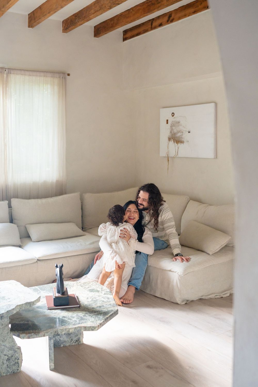 Family lounging together on a white couch in a bright room with wooden ceiling beams.
