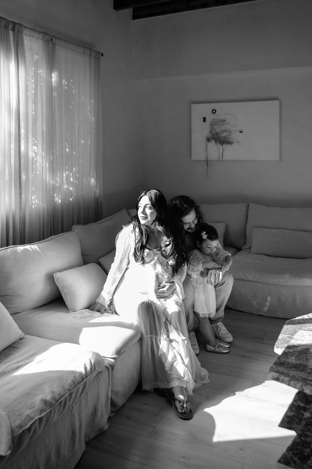 Sunlight streams through sheer curtains onto a white couch where a family shares an intimate moment.